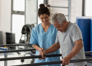 Physiotherapist helping a senior patient while he walks using his hands to support his weight on the bars and therapist looking very happy